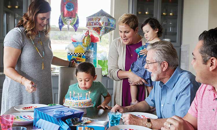 A family gathers around a table while a young boy blows out candles on a birthday cake