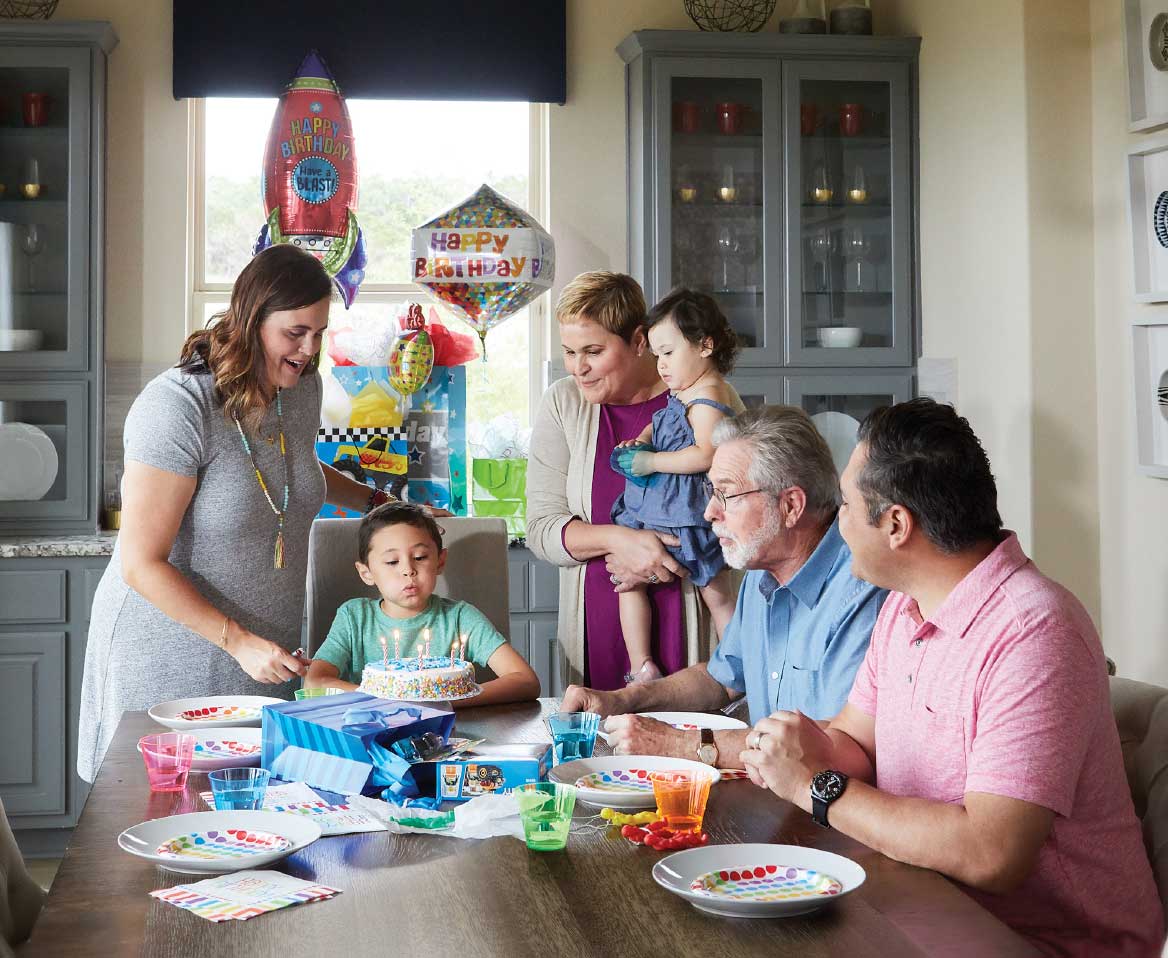 A multi-generational family sitting around a dining table with birthday balloons while a child blows out candles on a birthday cake