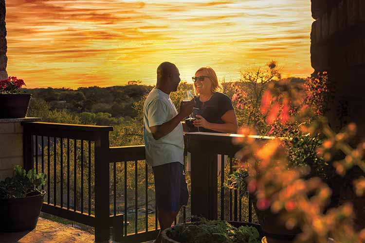 a couple enjoys glasses of wine while standing on a deck with the sunset behind them