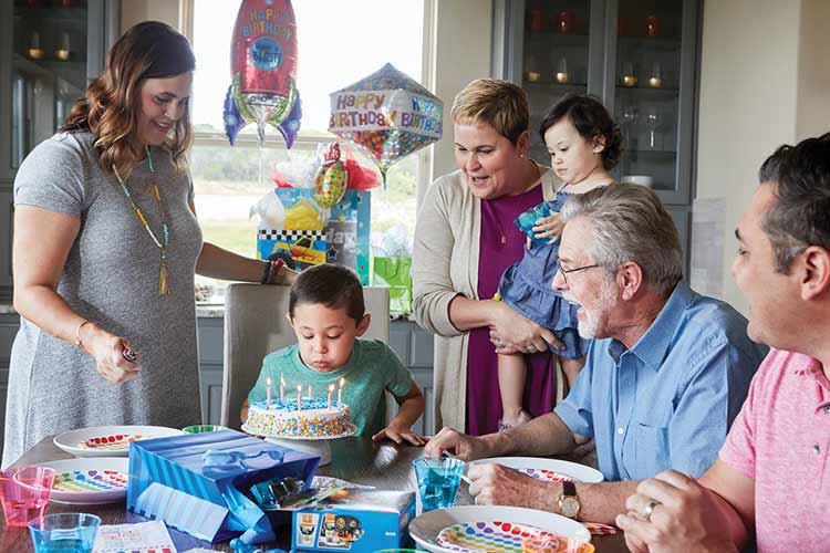 a family gathers at a table, a young boy blows out candles on a birthday cake