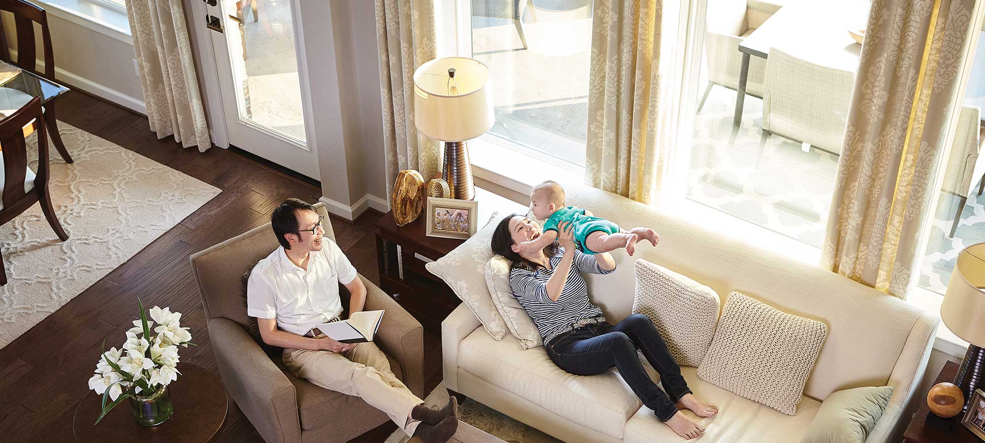 a man and woman sitting on couches by in a family room with large windows, the woman is holding a baby