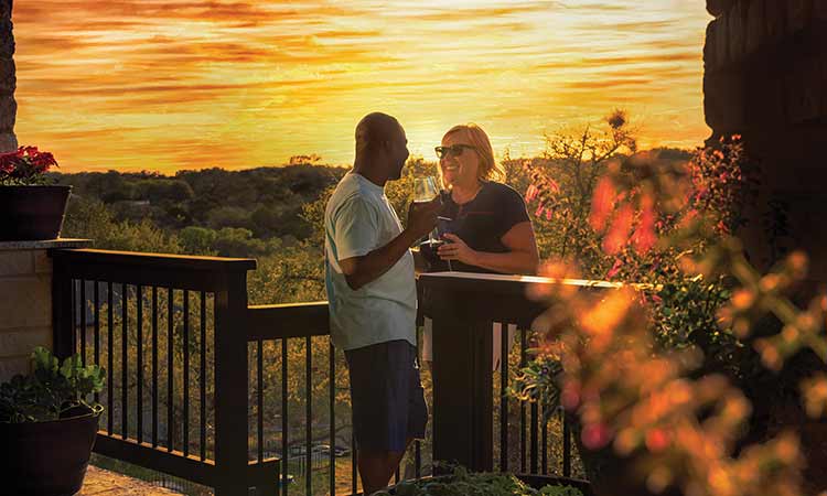 A man and a woman stand on a deck holding wine glasses, the sun is setting behind them