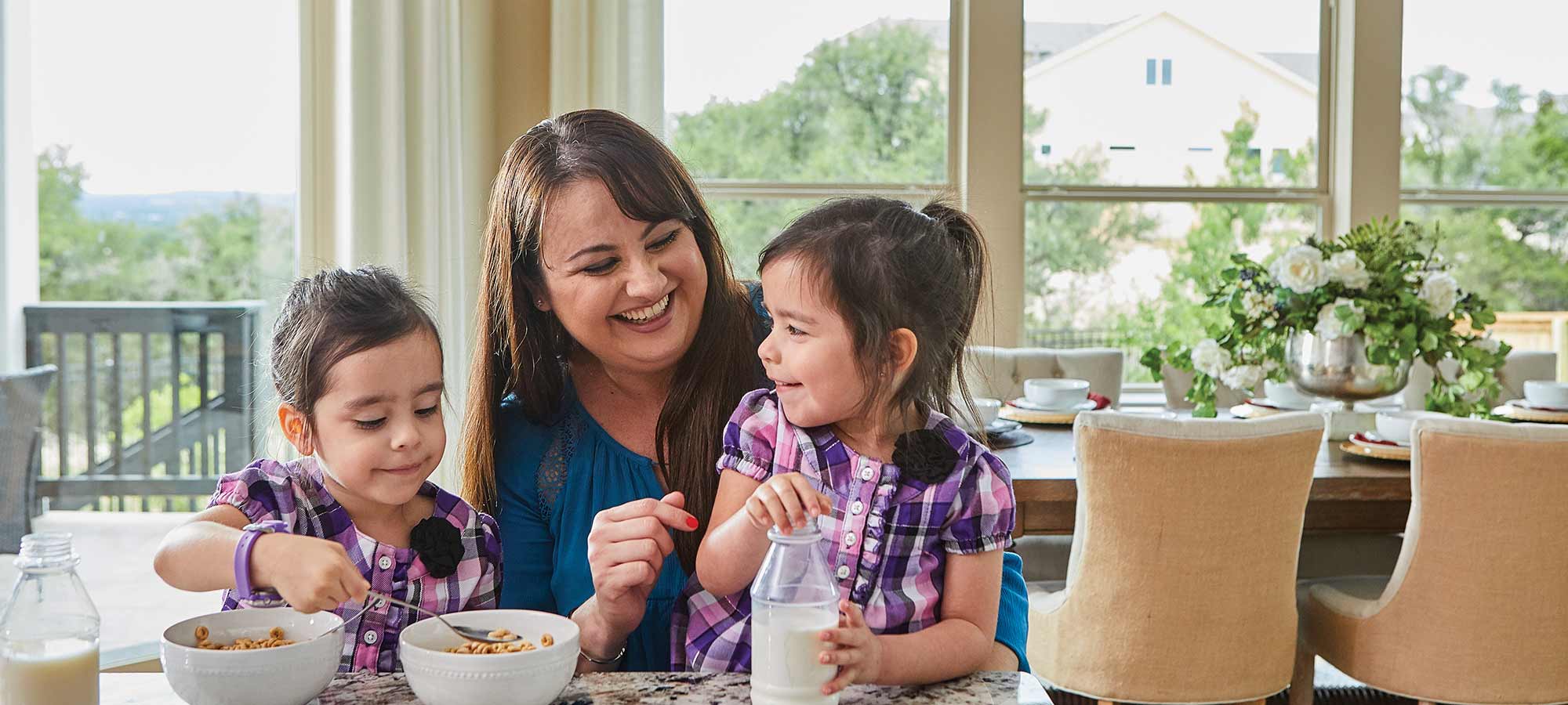 A woman sits at a kitchen counter with two young children who are eating from bowls of cereal