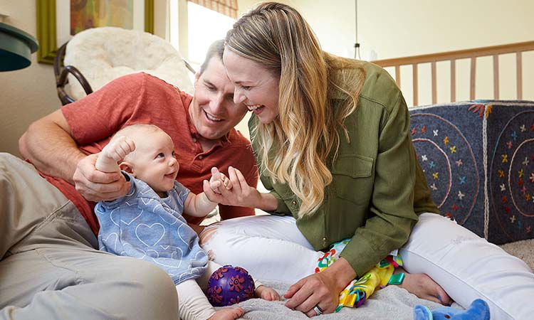 David Weekley Homes Homeowners - a man, woman and infant sitting on a bed with baby toys